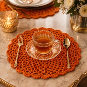 May include: A round, burnt orange crocheted placemat with a scalloped edge. Centered on the placemat is a clear, ribbed glass teacup and saucer filled with amber tea. A gold-toned fork is placed to the left and a gold-toned spoon to the right.