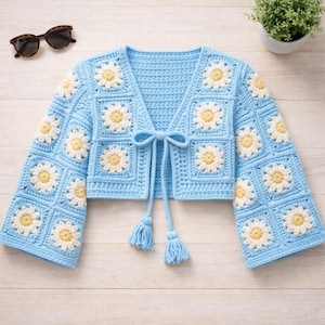 Può includere: Un cardigan all'uncinetto azzurro con motivi a margherita. Il cardigan ha maniche lunghe e una chiusura con fiocco e nappe. Gli occhiali da sole sono visibili in alto a sinistra.