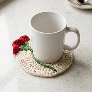 Puede incluir: Una taza de cerámica blanca lisa sobre un posavasos redondo de ganchillo color crema, con una flor roja con tallo y hojas verdes cosidas en la superficie. El posavasos es circular y presenta un patrón de hilo tejido con textura.