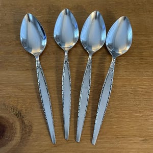 May include: Four silver-toned metal spoons arranged side by side. Each spoon features a smooth, polished oval bowl and a tapered handle with a textured, decorative border pattern along the edges, transitioning into a plain, matte centre panel.