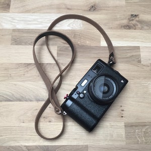 May include: A black digital camera with a brown leather strap rests on a wooden surface. The camera lens is central, and the strap is looped around the camera. The camera has a silver top and red button.