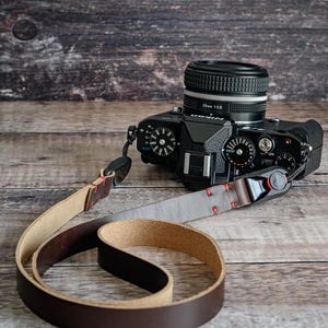 May include: A black Nikon camera with a 28mm 1:2.8 lens attached to a dark brown leather camera strap. The strap features a smooth dark brown exterior, a light tan suede interior, and is secured with red stitching and black plastic connectors.