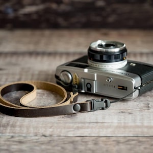 Puede incluir: Una cámara vintage plateada y negra con una correa de cuero. La cámara tiene una lente negra y una correa de cuero marrón con una hebilla negra. La cámara está sobre una superficie de madera.