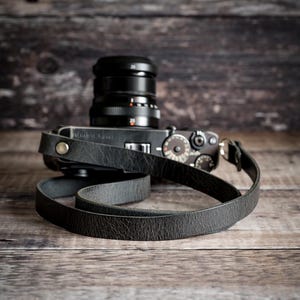 May include: A black camera with a lens and a leather strap rests on a wooden surface. The camera strap is looped and lies in front of the camera body. The background is a dark wooden wall.