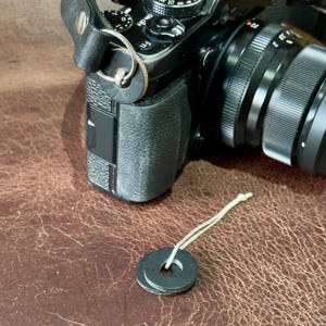 以下が含まれることがあります： 革製のストラップ、レンズ、コントロールダイヤルを備えた黒いデジタルカメラ。白い紐が付いた2つの黒いワッシャーが前景にあります。カメラは茶色の革の表面にあります。