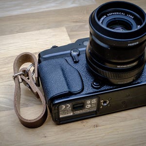 May include: Black digital camera with a lens attached, resting on a wooden surface. The lens is labeled "FUJINON ASPHERICAL LENS". A brown leather wrist strap with silver hardware is attached to the camera.
