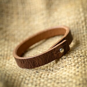 May include: A brown leather bracelet with a simple design. The bracelet has a loop and a metal stud fastening. The leather has a textured appearance. The bracelet is set against a neutral background.