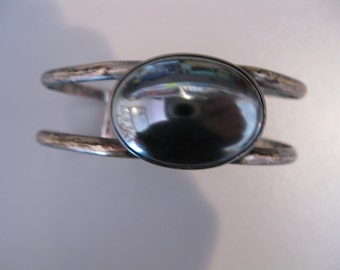 May include: A silver-toned cuff bracelet with a large, oval, dark gray stone at the center. The bracelet has a simple, open design with two parallel bands that curve around the wrist. The stone reflects light, creating a glossy appearance.