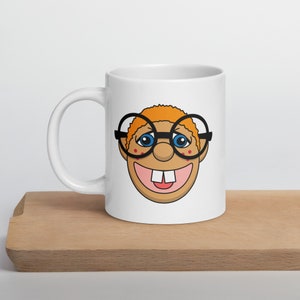 May include: White ceramic mug with a cartoon illustration of a smiling character with orange hair, blue eyes, and a toothy grin wearing black-rimmed glasses.