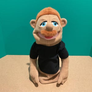 Op de afbeelding: Een pluchen pop met een lachend gezicht, rood haar en een bijpassende snor. De pop heeft blauwe ogen en draagt een zwart t-shirt. De huid van de pop is lichtbruin van kleur. De pop zit op een lichtbruin oppervlak tegen een turquoise achtergrond.