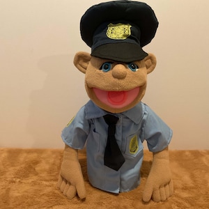 Puede incluir: Una marioneta de peluche vestida de policía. La marioneta tiene una cara bronceada, ojos azules y una boca ancha y sonriente. Lleva una camisa de uniforme azul con una corbata negra y un gorro de policía negro con una insignia dorada.