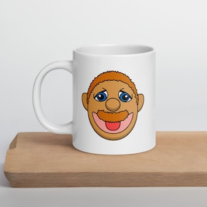 May include: White ceramic mug featuring a cartoon face with orange hair, a brown mustache, and blue eyes. The mug is on a wooden surface.