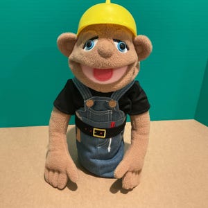 May include: A plush puppet with tan fleece skin, blue eyes, and a pink mouth, wearing a bright yellow plastic hard hat, a black short-sleeved shirt, and blue denim-style overalls with a black belt featuring a gold buckle and tool accents.