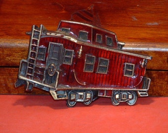 May include: A decorative metal caboose with a red body and gold-colored trim. The caboose features a ladder, windows, and wheels, and is set against a red surface and a wooden background.