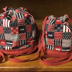 May include: Two red drawstring bags with red straps. The bags are made of a patchwork fabric with red, white, and blue stars and stripes. The bags have the word "USA" printed on them.