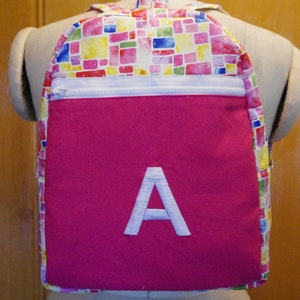 May include: A pink backpack with a white zipper and a white embroidered letter A on the front. The backpack has a colourful mosaic pattern on the top half.