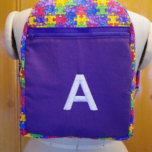 Puede incluir: Mochila morada con un bolsillo con cremallera y una letra A blanca bordada en la parte delantera. La mochila está hecha de una tela con un estampado de piezas de rompecabezas de colores.