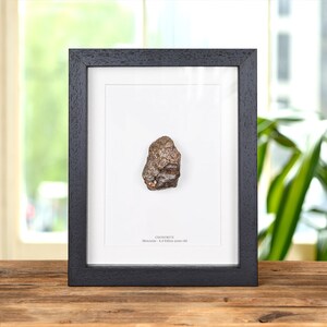 May include: A dark brown, textured chondrite meteorite fragment displayed in a black rectangular frame with a white mat. The text below the specimen reads: CHONDRITE Meteorite - 4.4 billion years old. The frame has a wood grain finish.