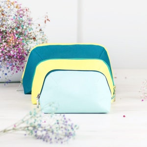 Puede incluir: Tres estuches con cremallera anidados en azul verdoso, amarillo y azul claro. Los estuches están hechos de un material suave de tela y tienen una forma curva.