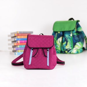 Op de afbeelding: Een gewatteerde magenta rugzak met zwarte randen en lichtblauwe ritstrekkers. Een tweede rugzak met een groene flap en een tropisch bladerpatroon is zichtbaar op de achtergrond. Een stapel kleurrijke boeken staat ook op de achtergrond.