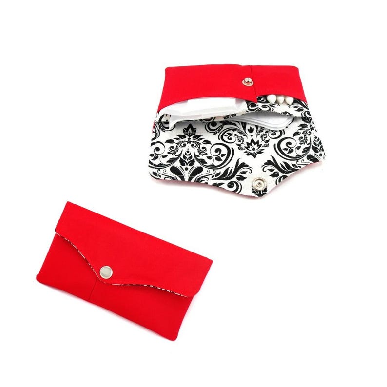 May include: A red fabric pouch with a snap closure. The pouch is open and shows a white fabric lining with a black and white damask pattern. The pouch is empty.