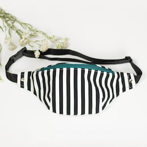 May include: A black and white striped fanny pack with a teal zipper and black adjustable strap. The bag is made of fabric and has a silver metal snap on each side. The background is white.