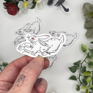 May include: White sticker featuring a black line drawing of a rabbit with red eyes, the words "good luck" are written below the rabbit. Several similar stickers are visible in the background, along with greenery.