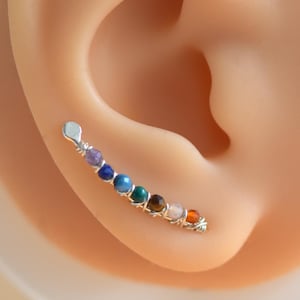 May include: A silver ear climber earring featuring a line of colourful gemstones. The gemstones include purple, blue, green, and orange. The earring is displayed on a model ear.
