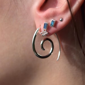 May include: A silver spiral hoop earring worn in the earlobe, accompanied by two smaller stud earrings featuring rectangular blue stones and a small metallic ball stud. The spiral earring has a polished finish and a curved, tapering design.