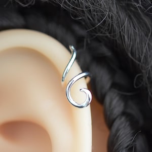 May include: A silver-colored ear cuff featuring a smooth, continuous wire design that curves into a spiral shape. The metal piece is positioned along the outer edge of the ear cartilage, showcasing a polished, reflective surface and a simple, open-ended structure.
