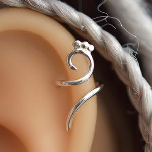 Op de afbeelding: Een zilveren oor manchet wikkelt zich om het oor, met een gebogen ontwerp met een kleine cluster van kralen aan de bovenkant. De sieraden worden getoond op een mannequin oor, met gevlochten haar op de achtergrond.