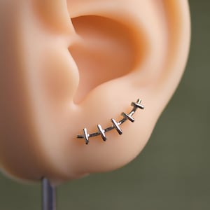 May include: A silver-coloured ear cuff earring shaped like stitches is shown on a mannequin ear. The earring curves along the ear's edge, with small, horizontal bars connected by vertical lines, resembling a stitched wound.