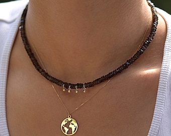 May include: A layered necklace set featuring a short strand of dark brown faceted beads with three small gold-toned dangling charms and a delicate gold-toned chain with a circular pendant depicting a cutout map of the world.