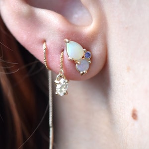 May include: A pair of gold earrings featuring a teardrop-shaped opal and a smaller blue gemstone. The earrings are attached to a thin gold chain that hangs down from the earlobe.