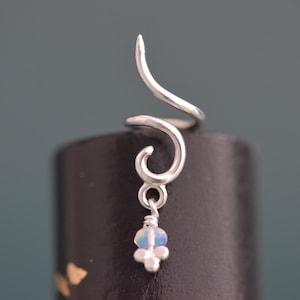 May include: A silver ear cuff featuring a spiralling, curved wire design that tapers to a point at the top. A small, dangling charm hangs from the lower loop, consisting of a single iridescent, translucent bead topped with a small silver metal accent.