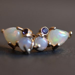 Puede incluir: Un anillo de tono dorado con dos &oacute;palos en forma de pera en los bordes exteriores, dos &oacute;palos redondos en el centro y dos peque&ntilde;as piedras preciosas redondas de color p&uacute;rpura entre los &oacute;palos. La banda de metal sujeta las piedras en racimo.