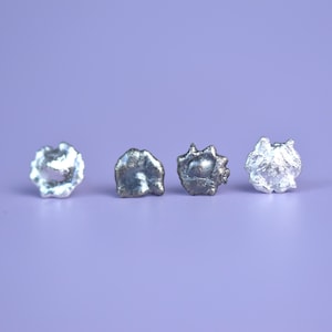 May include: Four small, irregularly shaped, metallic objects are arranged in a row against a solid purple background. The objects have a textured, possibly cast, appearance, with variations in colour from silver to darker grey.