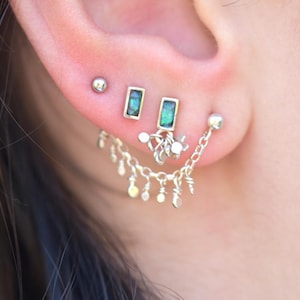 May include: A silver-toned ear piercing set featuring a small ball stud, two rectangular studs with iridescent green inlay, and a dangling chain connector with multiple tiny charms that drape across the lower lobe of the ear.