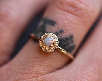 Könnte beinhalten: Ein Goldring mit einem runden Opal-Cabochon in der Mitte. Der Stein ist in eine kreisförmige Fassung mit einer texturierten, körnigen Oberfläche eingefasst. Die Ringschiene ist dünn, glatt und poliert, was einen Kontrast zur detaillierten Fassung bildet.