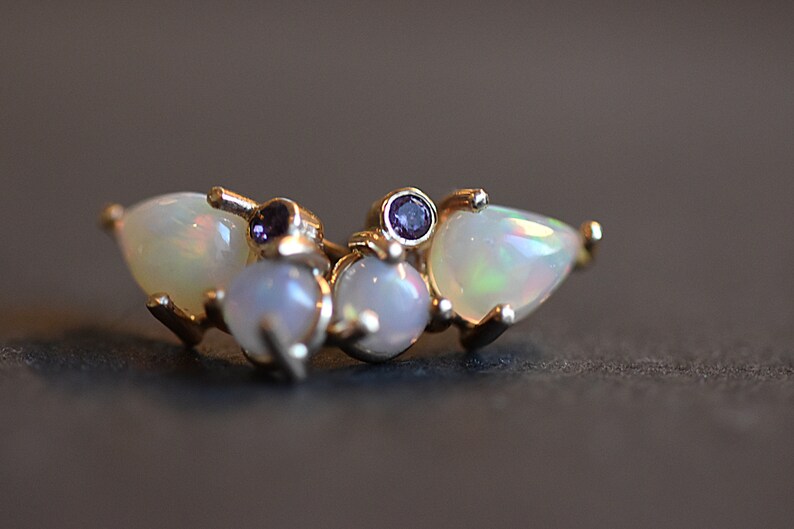 Puede incluir: Un anillo de tono dorado con dos &oacute;palos en forma de pera en los bordes exteriores, dos &oacute;palos redondos en el centro y dos peque&ntilde;as piedras preciosas redondas de color p&uacute;rpura entre los &oacute;palos. La banda de metal sujeta las piedras en racimo.