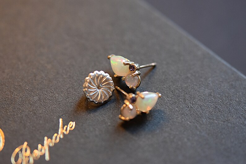 Puede incluir: Un par de pendientes de bot&oacute;n de plata con dos &oacute;palos en forma de l&aacute;grima y una peque&ntilde;a piedra preciosa morada en cada pieza. Los pendientes incluyen cierres met&aacute;licos circulares y texturizados. La palabra Bespoke se ve en letras doradas.