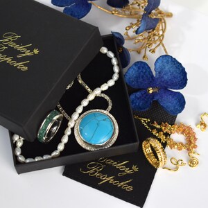 Puede incluir: Un joyero negro con el texto dorado Bailey Bespoke que contiene un collar de perlas blancas, un colgante de piedra turquesa y un anillo con incrustaciones verdes. Junto a la caja hay anillos dorados, una cadena y una bolsa de tela negra.