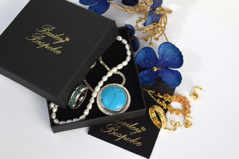 Puede incluir: Un joyero negro con el texto dorado Bailey Bespoke que contiene un collar de perlas blancas, un colgante de piedra turquesa y un anillo con incrustaciones verdes. Junto a la caja hay anillos dorados, una cadena y una bolsa de tela negra.