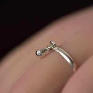 Puede incluir: Primer plano de un anillo plateado con un diseño abstracto. El anillo presenta un elemento en forma de lágrima y un pequeño lazo. El anillo se lleva en un dedo, con un fondo borroso.