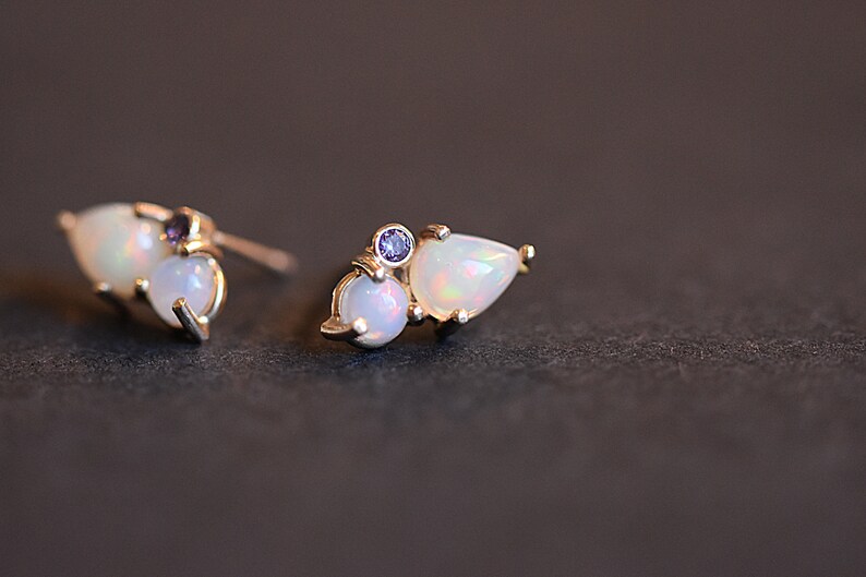 Puede incluir: Un par de pendientes de bot&oacute;n con un &oacute;palo en forma de l&aacute;grima y un &oacute;palo redondo engastados en metal dorado, acentuados con una peque&ntilde;a piedra preciosa redonda de color p&uacute;rpura. Los pendientes muestran destellos iridiscentes en las piedras.