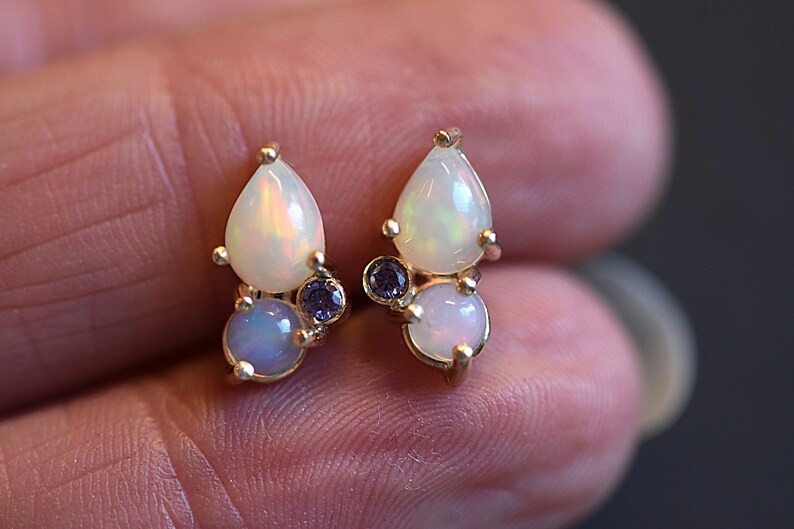 Puede incluir: Un par de pendientes de bot&oacute;n con engaste dorado, una &oacute;pala en forma de pera, una &oacute;pala redonda y una peque&ntilde;a piedra preciosa redonda de color p&uacute;rpura. Cada pendiente muestra destellos iridiscentes en las &oacute;palas blancas y un tono violeta.