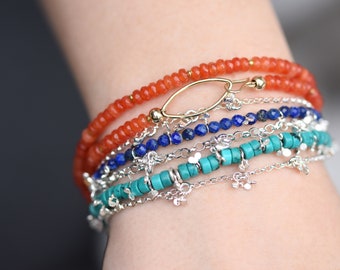 Puede incluir: Una pila de cinco pulseras que incluye una hilera de cuentas naranjas facetadas con un cierre ovalado dorado, una hilera de cuentas azules de lapislázuli y tres cadenas plateadas intercaladas con cuentas cilíndricas turquesas y pequeños dijes.