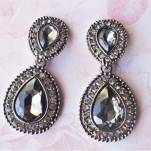 May include: A pair of dangle earrings with two teardrop-shaped crystals. The crystals are clear with a slight gray tint and are surrounded by smaller clear crystals. The earrings are silver-toned.