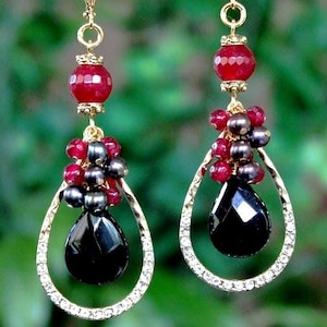 May include: A pair of elegant dangle earrings. Each earring features a faceted black teardrop-shaped gem, surrounded by a gold-colored frame encrusted with small clear crystals. Above the teardrop are clusters of red and dark gray beads, and a large red bead at the top.