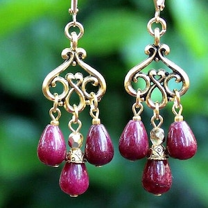 May include: A pair of drop earrings featuring an antiqued gold-tone metal filigree top piece with a heart-shaped cutout. Three deep magenta, teardrop-shaped beads hang from each filigree component, connected by small gold-tone metal jump rings.
